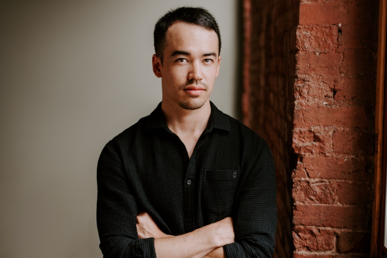 David's headshot - he wears a black collared shirt and has his arms crossed while he stares at the camera. the background is half a brick wall and half a neutral toned wall.